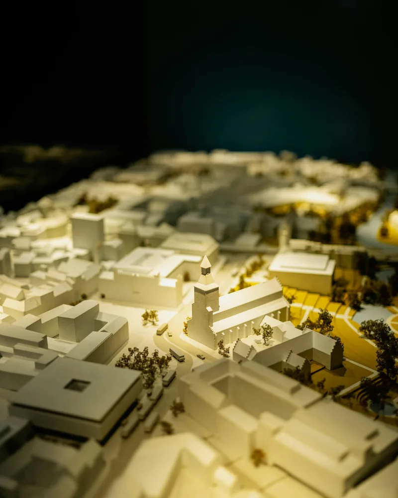 Maqueta urbana blanca iluminada desde el interior, con calles marcadas por luz cálida y arbolado en miniatura
